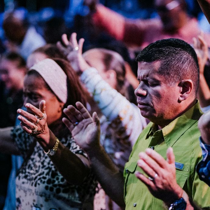 A vibrant crowd of diverse adults worshipping with raised hands indoors.