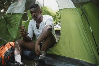 Sad African American male traveler resting in tent and browsing Internet on cellphone while spending journey in nature